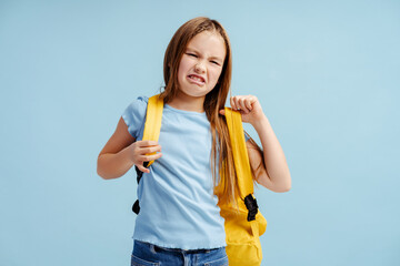 Upset crying little girl, pupil wearing casual clothes backpack isolated on blue background