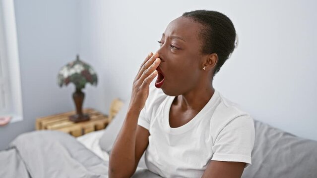 Exhausted african american woman lying bored in bed, yawning tiredly, sleepiness washing over her, hand covering her yawning mouth, struggling with sleep in her comfortable bedroom