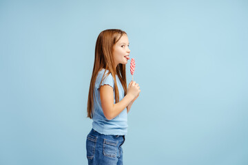 Portrait of positive little girl wearing casual clothes, holding lollipop, looking at it, eating