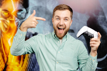 Young bearded man standing at the wall in the room and playing, holding wireless joystick,...