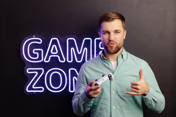 Young bearded man standing at the wall in the room and playing, holding wireless joystick, screaming, and winning intense video game match