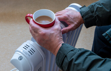 A person holding a hot cup of tea and warming the hands on a portable oil filled heater electric...