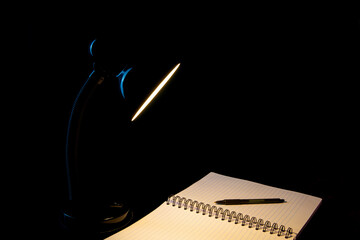 Black Desk Lamp Shining on a Lined Pad an Pen on a Black Background - 690204858