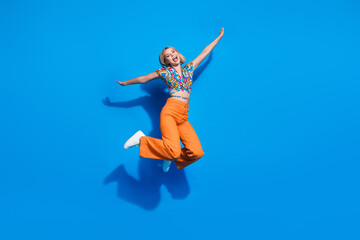 Full size photo of cute good mood girl wear blouse orange trousers jumping holding palms like wings isolated on blue color background