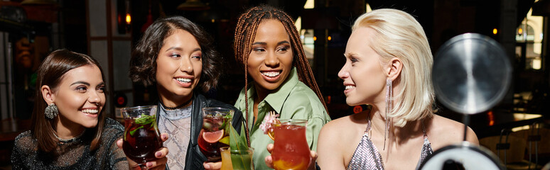 smiling and elegant multicultural female friends holding cocktails glasses in bar, horizontal banner