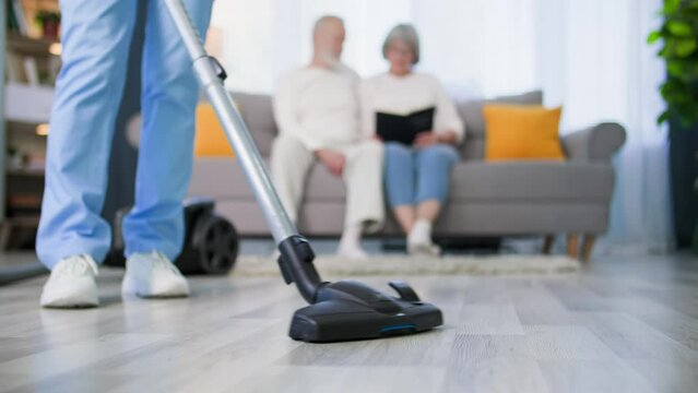 caring about elderly people, social worker in a medical uniform with a vacuum cleaner in her hands helps an elderly couple with cleaning house, close-up
