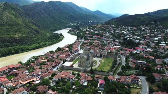 Aerial perspective of Mtskheta, Georgia, showcasing the renowned Svetitskhoveli Cathedral. The city unfolds along the riverbank, surrounded by mountains, creating a picturesque scene from above.