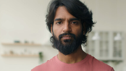 Close up front view adult 30s multiracial Arabian Indian ethnic bearded man serious looking at camera. Male head shot portrait of calm middle-aged muslim guy homeowner home renter posing alone indoors