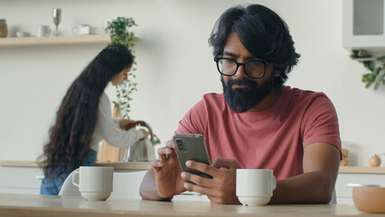 Bearded muslim Indian Arabian man remote working from home kitchen use mobile phone apps cellphone...