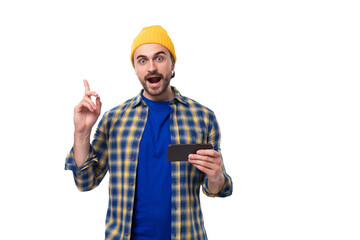 young stylish brunette man with a brutal beard and mustache dressed in a hat and shirt is inspired by the idea holding a smartphone