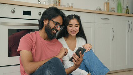 Arabian Indian interracial couple woman man sit on warm floor in kitchen watching funny video...