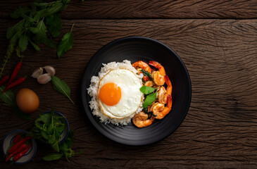 Stir-fried shrimp with basil Thai street food arranged on a black plate with a fried egg Spicy Thai food placed on a wooden table. Top view.