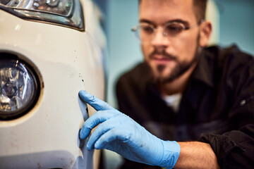 A male mechanic notices a scratch on the white car, using gloves.