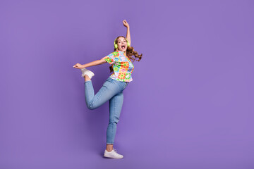 Full size photo of funky girl dressed print t-shirt jeans in headphones dancing on one leg hand up isolated on violet color background