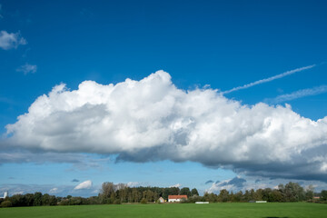 Fototapeta na wymiar This image presents a breathtaking view of expansive blue skies filled with billowing white clouds over a serene rural landscape. A vast green field stretches towards a distant horizon, where a small