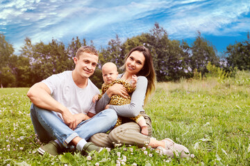 Young parents in their 20s with six month old baby are sitting on grass of backyard lawn of country house.