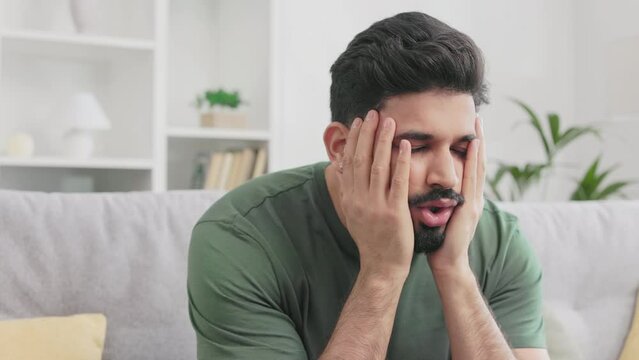 Stressed hindu guy touching head and gesturing from frustration while sitting on grey couch at living room. Bearded male person in casual wear experiencing negative emotions at home.