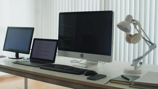 Medium close up of part of workplace in programmers office, table with open laptop, computers with codes on screens and smartphone