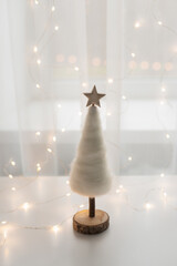 Tender white Chhristmas tree with wooden star in a backlight of the window with fairylights. Festive holidays concept.