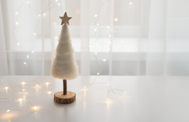 Tender white Chhristmas tree with wooden star in a backlight of the window with fairylights. Festive holidays conbcept, copy space for your text.