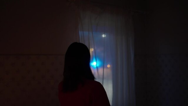 Woman Looks Out The Window Of Her House Observing Night Incident Scene With Out Of Focus Police Lights