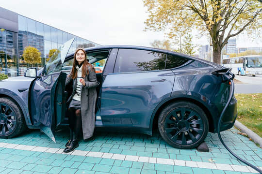 Young positive woman in grey coat with cup of coffee waiting while her electric car charging, sustainable and economic transportation concept.