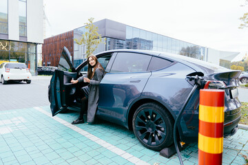 Young business woman in grey coat with cup of coffee waiting while her electric car charging,...