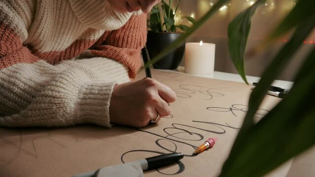The girl is painting the text. Writes the text on a large kraft paper. Decoration for home. Evening time. In the background there are green plants and a candle