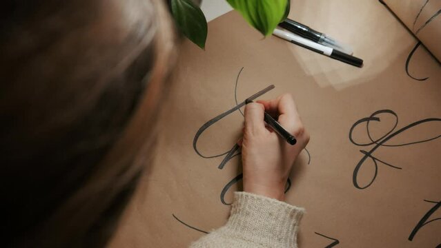 The girl is painting the text. Writes the text on a large kraft paper. Decoration for home. Evening time. In the foreground is a green plant. Top view.