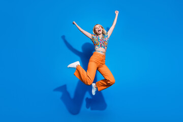 Fototapeta na wymiar Full size photo of ecstatic optimistic girl wear blouse orange trousers jumping hands up win betting isolated on blue color background