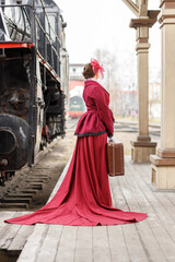 A beautiful girl in a burgundy suit of the last century and a hat with a veil stands with a suitcase in her hands near an old steam locomotive.Vintage portrait of the last century, retro journey