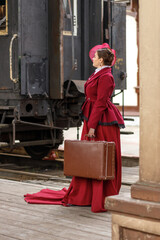 A beautiful girl in a burgundy suit of the last century and a hat with a veil stands with a...