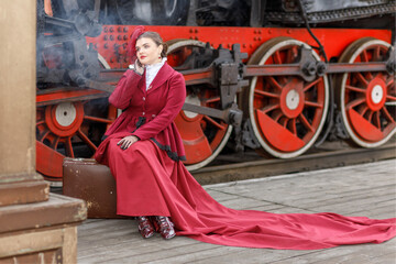 A beautiful girl in a burgundy suit of the last century and a hat with a veil is sitting on a suitcase near an old steam locomotive.Vintage portrait of the last century, retro journey