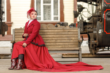 A beautiful girl in a burgundy suit of the last century and a hat with a veil is sitting on a suitcase near an old steam locomotive.Vintage portrait of the last century, retro journey