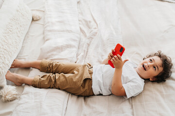 Playful curly Brazilian boy in white t-shirt and pants laying on bed laughing holding phone with...