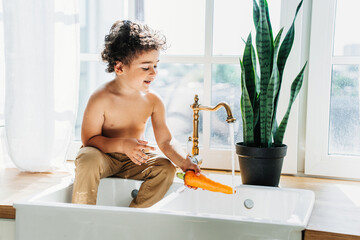Happy caucasian curly boy sitting on sink at kitchen washing carrot under water. Handsome Spanish kid at home helping to cook, cleaning vegetables. Healthy food concept, kids life. Healthy lifestyle.
