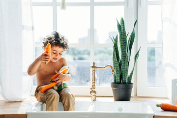 Little handsome curly caucasian boy sitting on sink at kitchen holding vegetables for wash them....