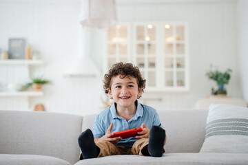 Awesome curly Brazilian boy laughing sitting on cozy sofa at home holding phone, playing games over...