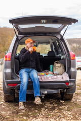 A Caucasian man sits in the trunk of a car and drinks coffee. Outdoor picnic. Road trip