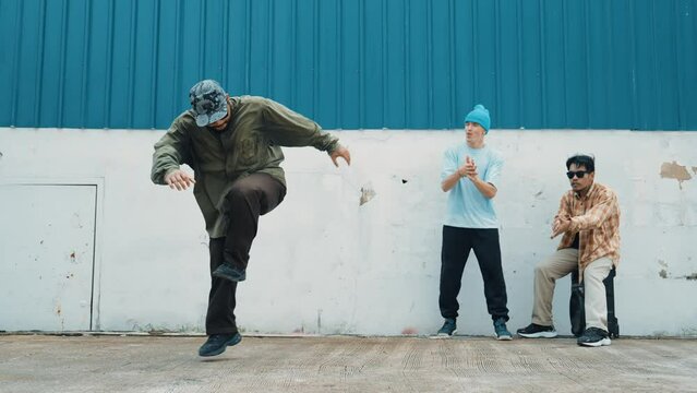 Skilled hipster doing cart wheel while multicultural friends encourage him. Hip-hop dancer moving foot step to music surrounded by group of hipster at wall near street. Outdoor sport 2024. hiphop.