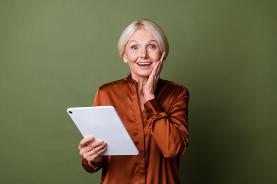 Photo portrait of pretty retired female hold tablet hand cheek shocked dressed stylish brown silk outfit isolated on khaki color background - Powered by Adobe