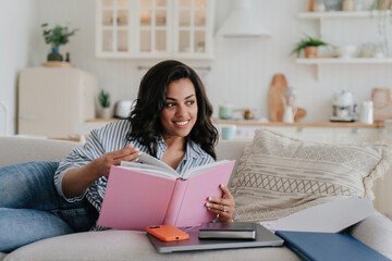 Cheerful African American young woman in striped shirt and blue jeans laying on sofa with opened book toothy smiling looks aside. Brazilian girl with wavy hair reading book against blurry kitchen.