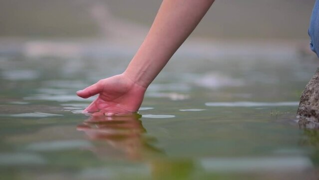 Slow motion video of a young woman taking her hand out of a lagoon of glacier water, with her nails painted purple and drops of water running down her fingers.