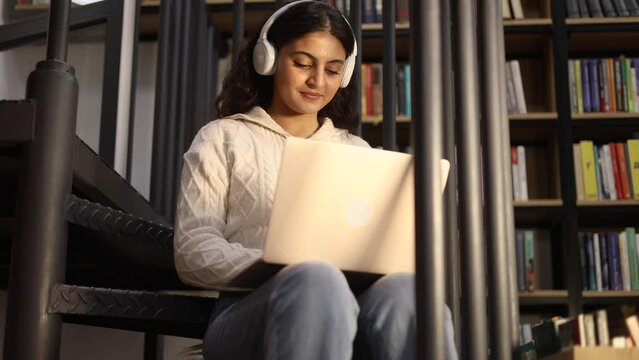 Portrait of pretty young woman student with headphones listening music while having distance remote education or work typing browsing scrolling on laptop computer at library or book store