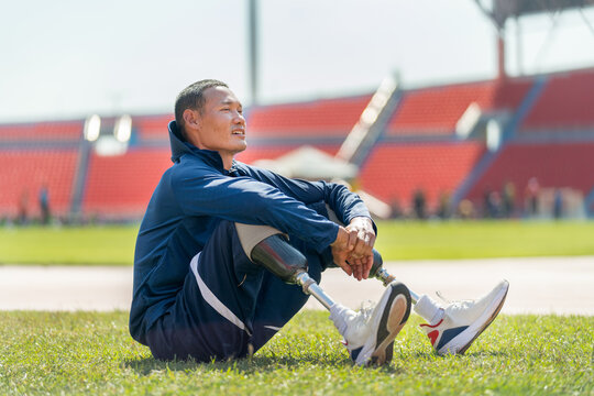 Sport man athlete with a prosthesis on his leg sit on grass field near the track of stadium to relax after exercise with day light.