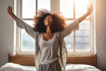 African American woman wakes up in the morning with her arms stretched out in the bedroom against the morning light. - obrazy, fototapety, plakaty