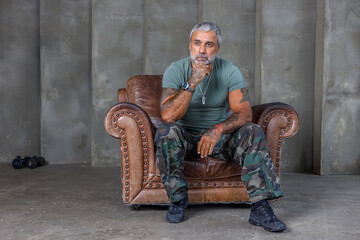 Upset young man in camouflage uniform soldier sitting at armchair over white studio background, looking at copy space, holding hands clasped, experiencing posttraumatic stress disorder, panorama