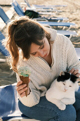 Happy young woman eating ice cream and cuddling a cat on the beach 