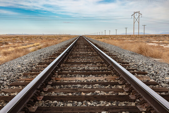 Closeup of railroad tracks in central Utah. Stretching straight to the horizon. Telephone poles to the right. Blue cloudy sky beyond.
