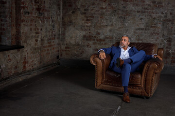 A handsome middle-aged man in a blue formal suit poses sitting on a leather armchair. Portrait of a...